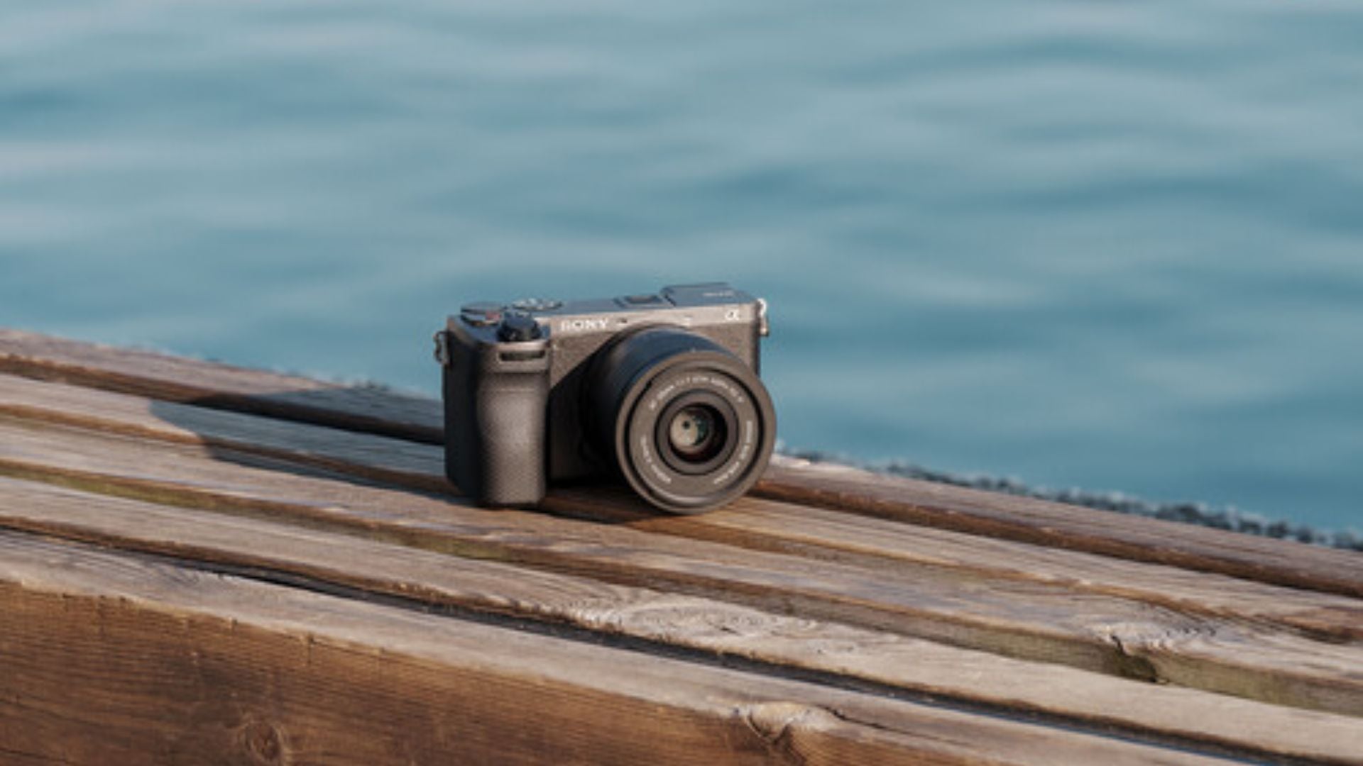 Gray Sony mirrorless camera with attached lens resting on rustic wooden planks with blurred blue water background in outdoor setting.