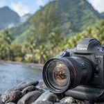 Professional Canon EOS-1D X Mark III camera with zoom lens positioned on rocky shore overlooking mountain lake with forested hills in background.