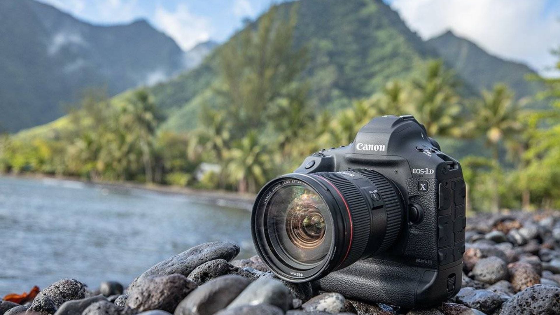 Professional Canon EOS-1D X Mark III camera with zoom lens positioned on rocky shore overlooking mountain lake with forested hills in background.