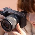 Smiling woman with reddish hair holding black Sony mirrorless camera with silver lens up to take photograph against warm peach background.