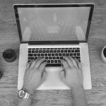 Black and white aerial shot of hands typing on MacBook Pro with DSLR camera, lens, wristwatch, and coffee cup arranged on wooden desk.