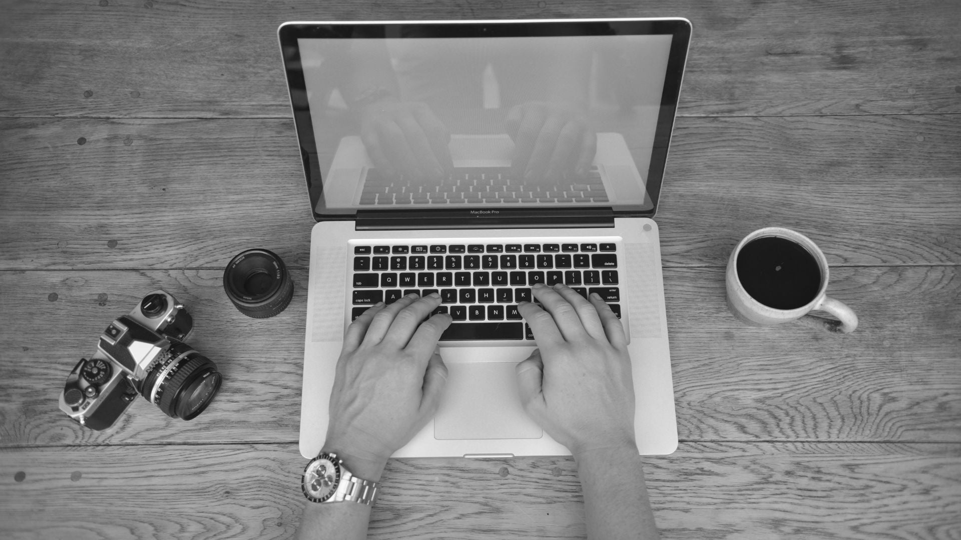 Black and white aerial shot of hands typing on MacBook Pro with DSLR camera, lens, wristwatch, and coffee cup arranged on wooden desk.