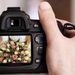 Hand holding black DSLR camera with LCD screen displaying captured image of pink and white rose bouquet with baby's breath and greenery.