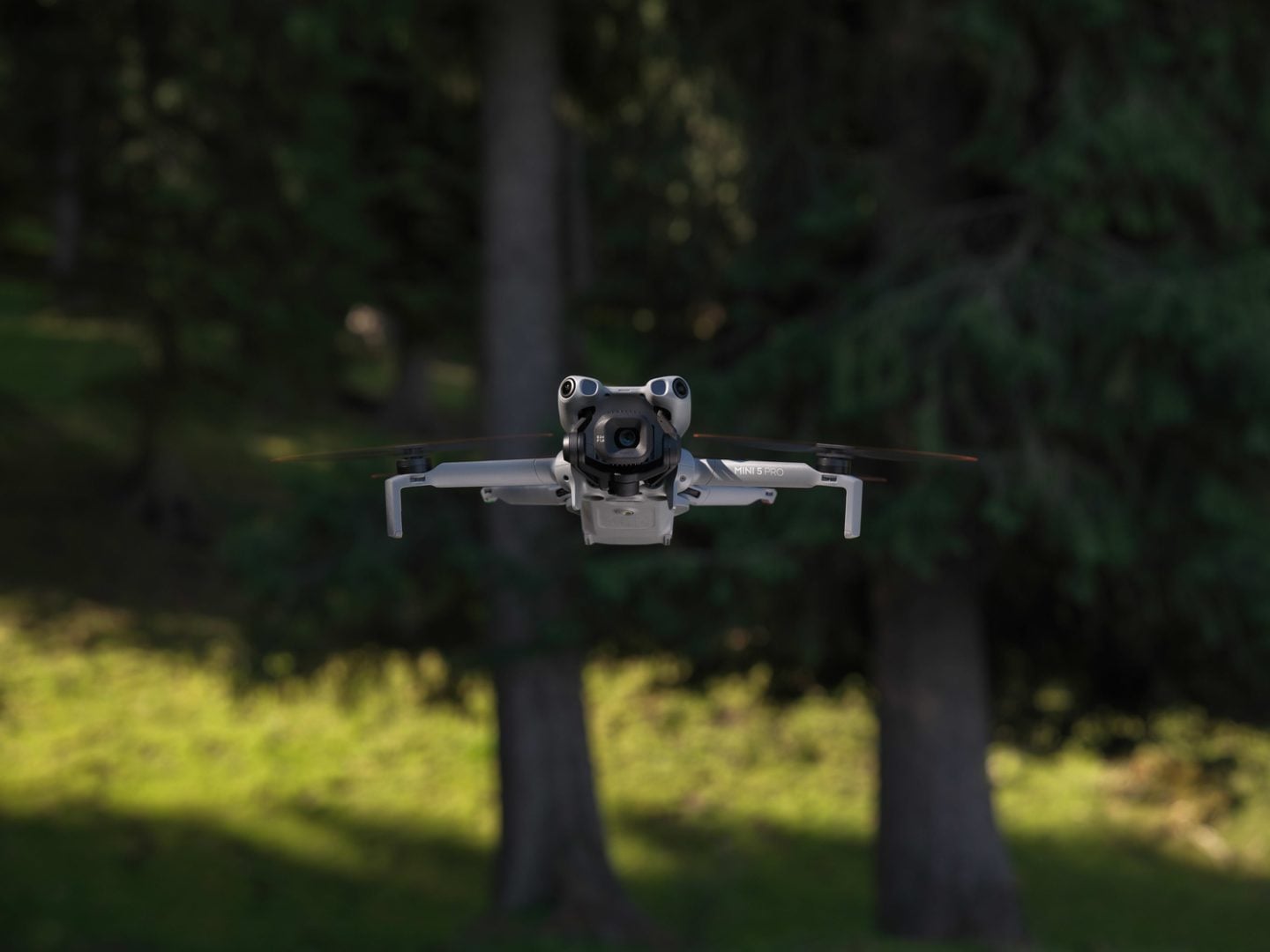 Gray DJI Mini 5 Pro drone with prominent camera gimbal flying against blurred green forest background with tree trunks and foliage.