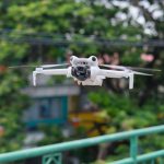 Compact white DJI Mini 5 Pro quadcopter with camera gimbal flying over lush green vegetation with blurred residential buildings in background.