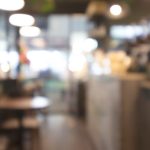 Soft out of focus view of modern restaurant or cafe interior with bright pendant lights, wooden tables, and people dining in warm ambient atmosphere.