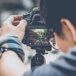 Hand adjusting DSLR camera mounted on tripod displaying live view screen showing outdoor scene with camera settings visible including aperture f/22 ISO 10 shutter 1/55s.