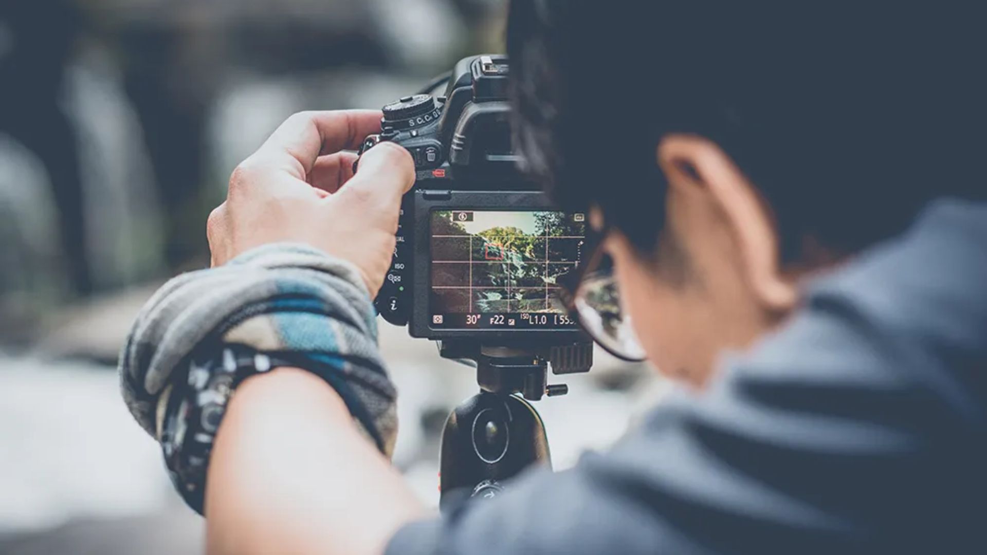 Hand adjusting DSLR camera mounted on tripod displaying live view screen showing outdoor scene with camera settings visible including aperture f/22 ISO 10 shutter 1/55s.