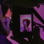 Filmmaker captures content creator on camera screen during professional video shoot with dramatic purple and pink lighting in modern studio setup.