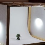 Homemade photo light box with white backdrop and two desk lamps illuminating small toy figurine for product photography on table.