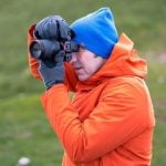 Photographer wearing bright orange jacket and blue beanie holding DSLR camera up to eye capturing photos outdoors in natural green blurred background setting.