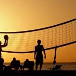 Players silhouetted against golden orange sunset sky during beach volleyball game with net stretched across frame and ocean in background.