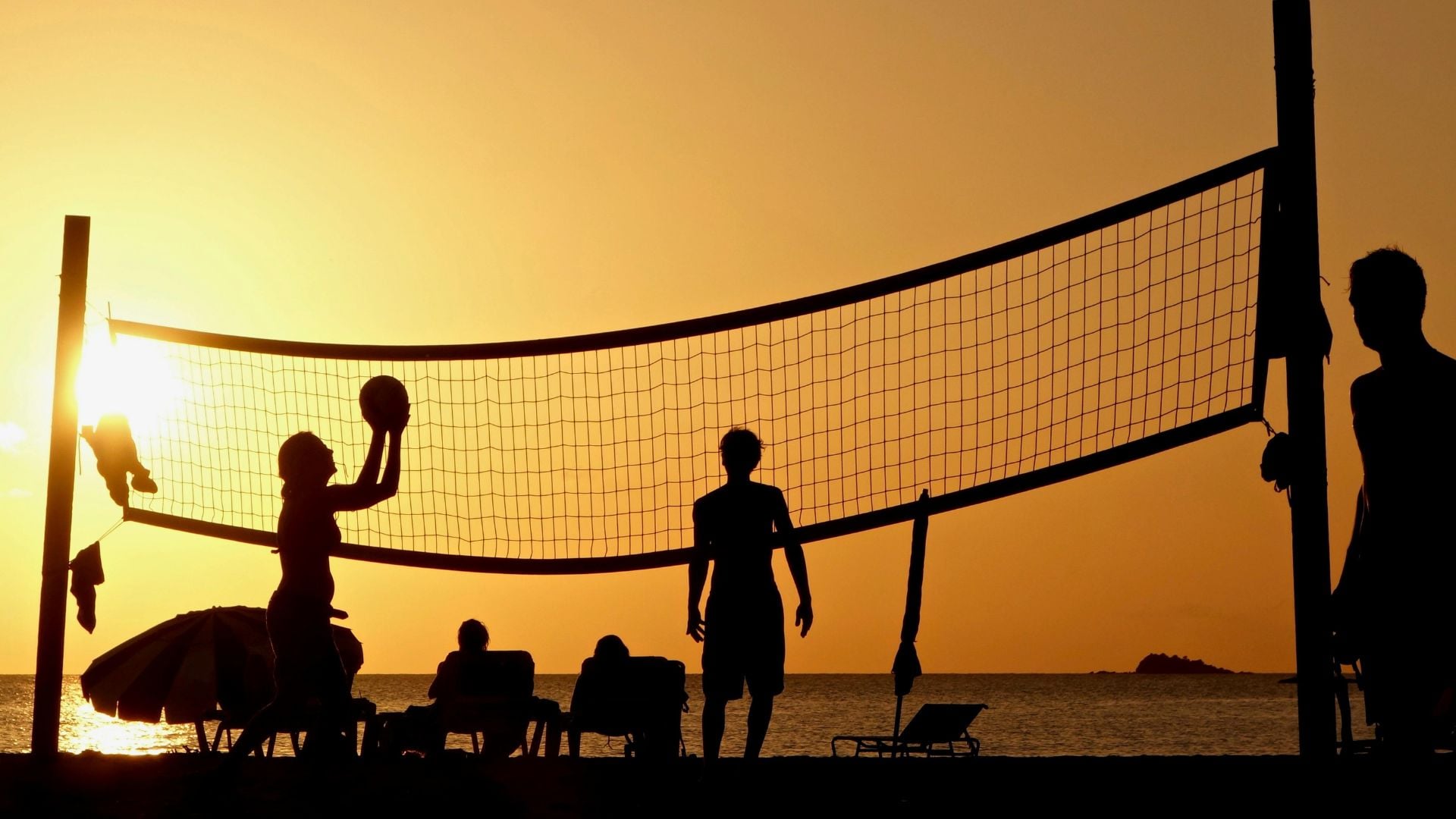 Players silhouetted against golden orange sunset sky during beach volleyball game with net stretched across frame and ocean in background.