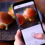 Hand holds smartphone photographing gourmet burger sliders on woven black placemat with camera interface visible on phone screen display.