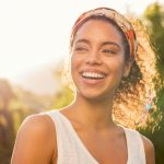 Smiling woman wearing orange headband and white tank top enjoying golden hour sunlight outdoors with natural greenery in blurred background creating warm atmosphere.
