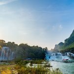 Multiple waterfalls cascading into turquoise river with tourist boat, dramatic limestone mountains backdrop, golden sunrise light creating misty atmospheric scene with lush greenery.
