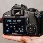 Hand holding Canon DSLR showing rear LCD screen with camera settings displayed: 1/100 shutter speed, F00 aperture, ISO 800, manual mode visible.
