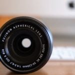 Close-up of black Fujinon aspherical lens showing front element with white text markings, wooden table surface and camera body blurred in background.