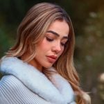 Woman with long blonde highlighted hair looking down wearing gray ribbed sweater with white fur collar trim outdoors with soft blurred natural background.