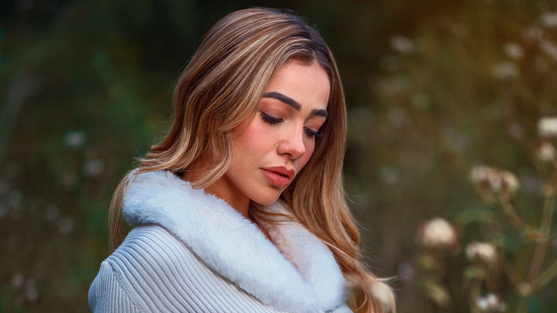 Woman with long blonde highlighted hair looking down wearing gray ribbed sweater with white fur collar trim outdoors with soft blurred natural background.
