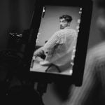 Black and white image showing camera monitor displaying man in striped shirt during photo shoot with photography equipment visible in dark studio setting.