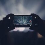 Hands holding smartphone recording live concert performance with dramatic stage lights and blue laser beams radiating from stage in dark venue.
