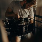 Man in white shirt and glasses operates professional cinema camera with mounted monitor indoors, focusing intently on equipment in dimly lit production environment.