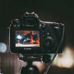 Canon DSLR on tripod displaying nighttime street scene with orange lights on LCD screen, bokeh lights visible in dark background creating atmospheric effect.