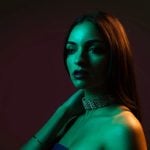 Woman in sequined collar top photographed with dramatic green and orange gel lighting against dark background creating modern editorial fashion photography style.