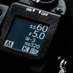 Close-up of camera LCD display showing shutter speed 600, aperture F5.0, ISO 320 settings with mode dial and on/off switch visible on black camera body.