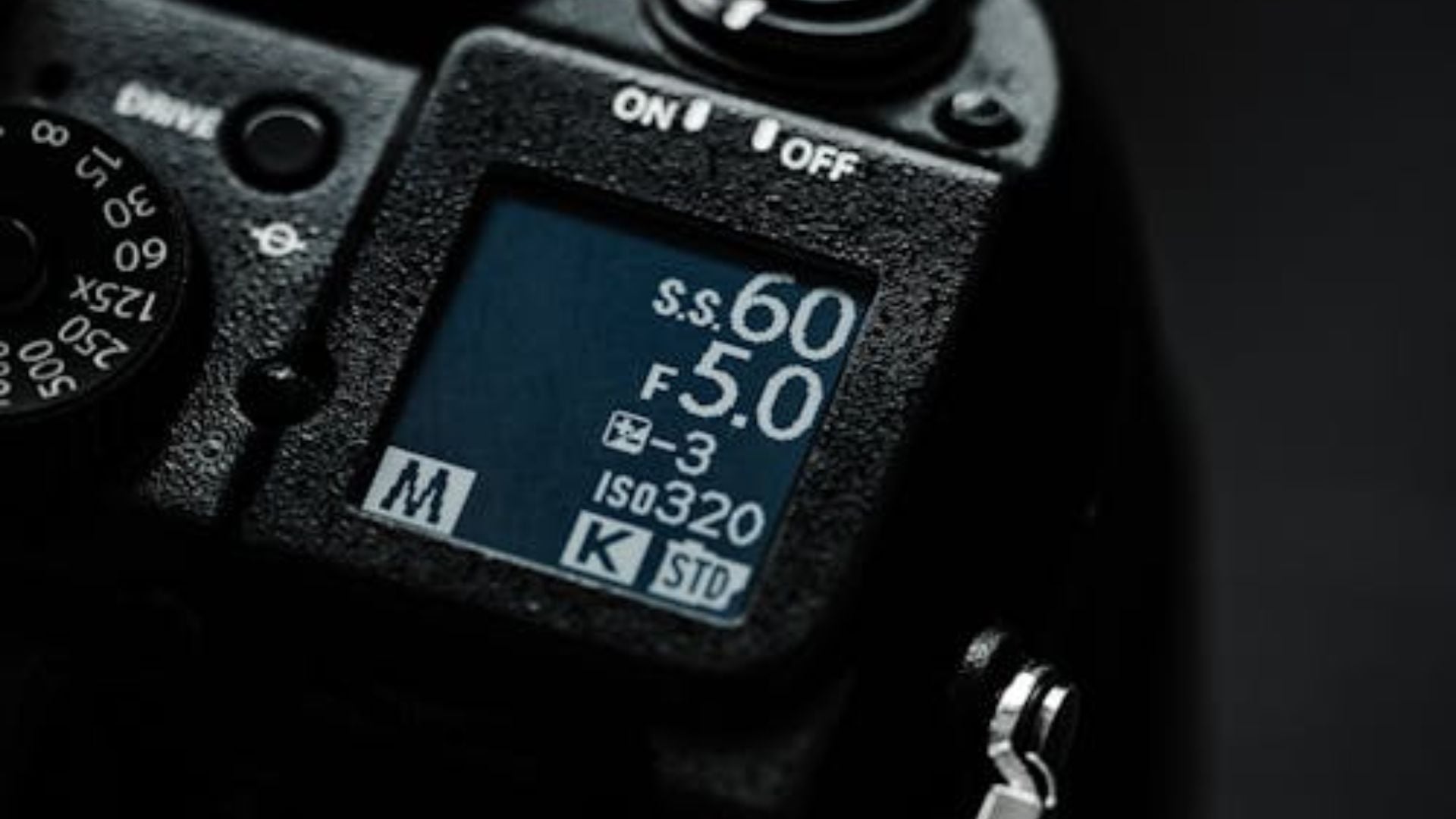 Close-up of camera LCD display showing shutter speed 600, aperture F5.0, ISO 320 settings with mode dial and on/off switch visible on black camera body.