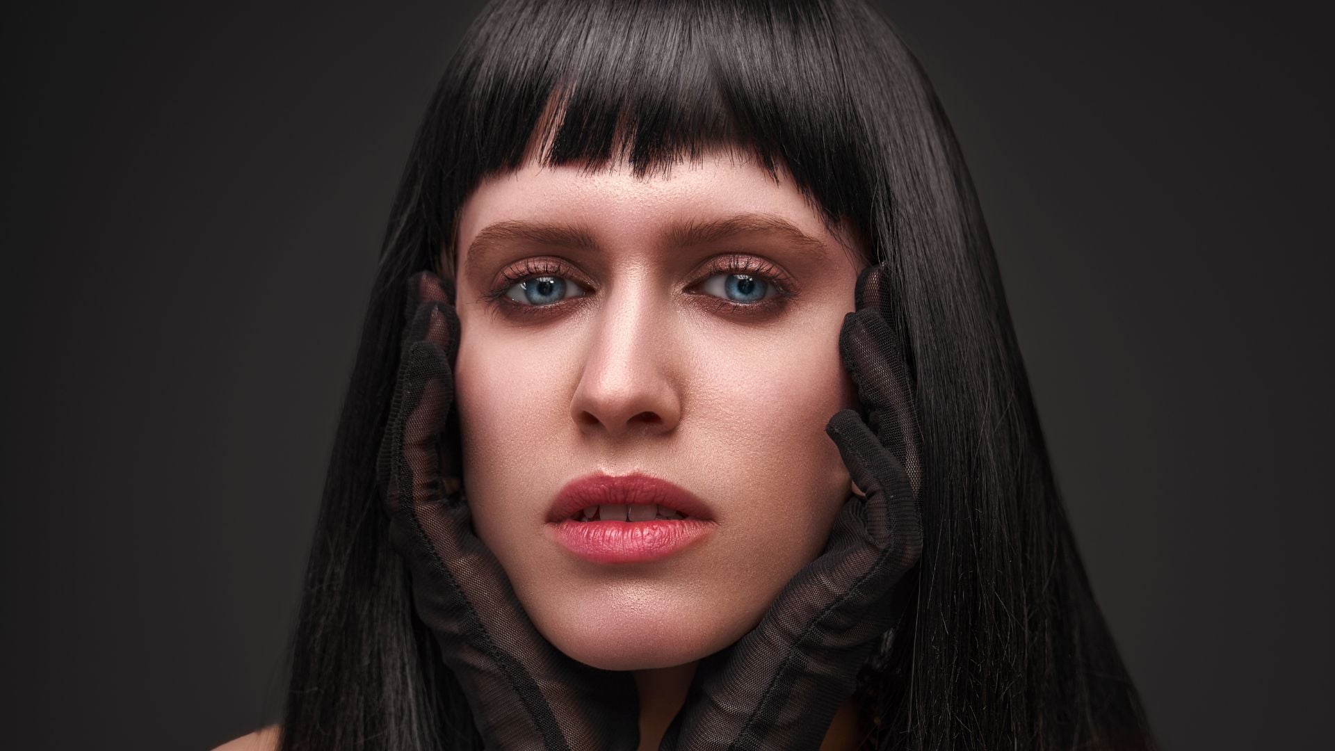 Woman with straight black hair and blunt bangs wearing sheer black gloves framing face, blue eyes with natural makeup against gray studio background portrait.