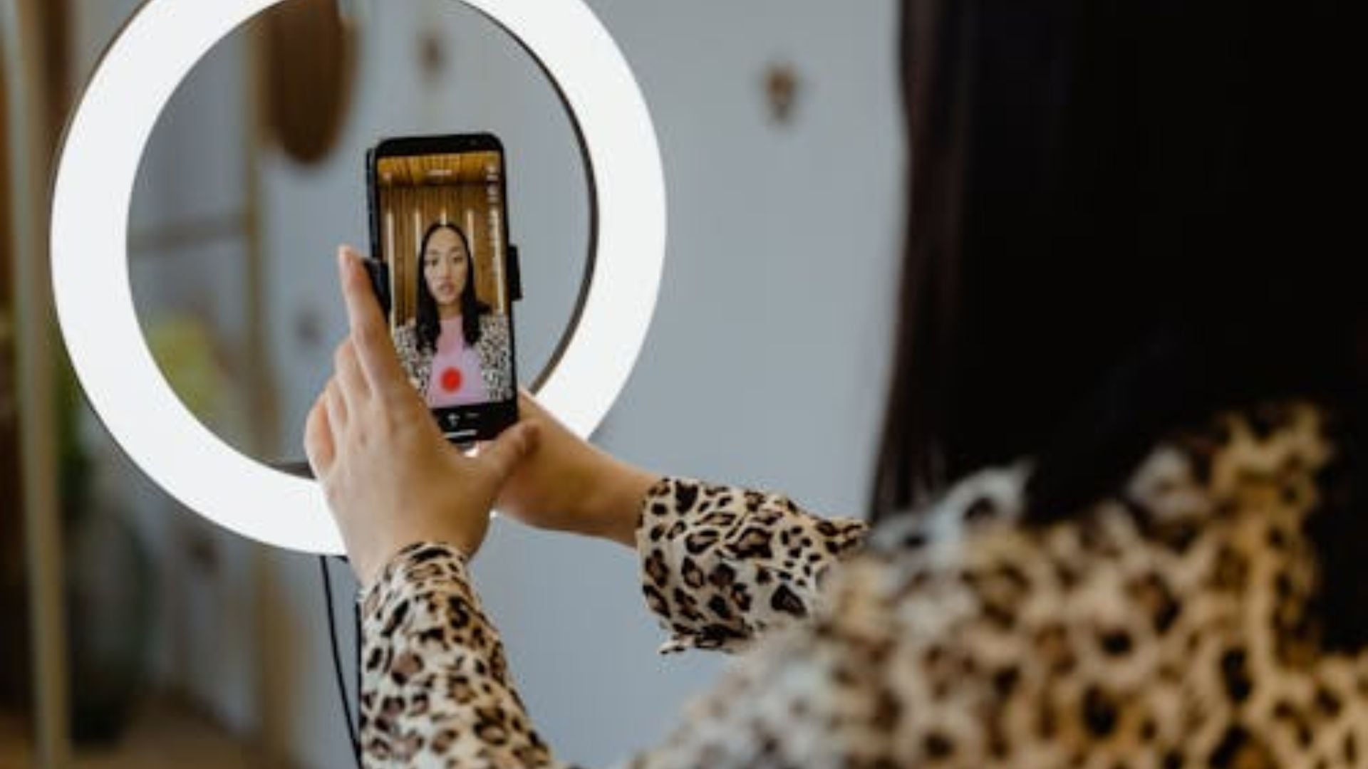 Person in leopard print sleeve holds smartphone mounted in LED ring light, recording video content with woman visible on screen in home.