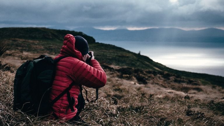 Person wearing red puffer jacket and black beanie crouching with camera photographing dramatic overcast coastal hillside with dark storm clouds and ocean view backdrop.