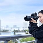 Asian woman in black outfit with ponytail holding DSLR camera photographing urban skyline with tall buildings across waterfront from elevated viewpoint with metal railing.