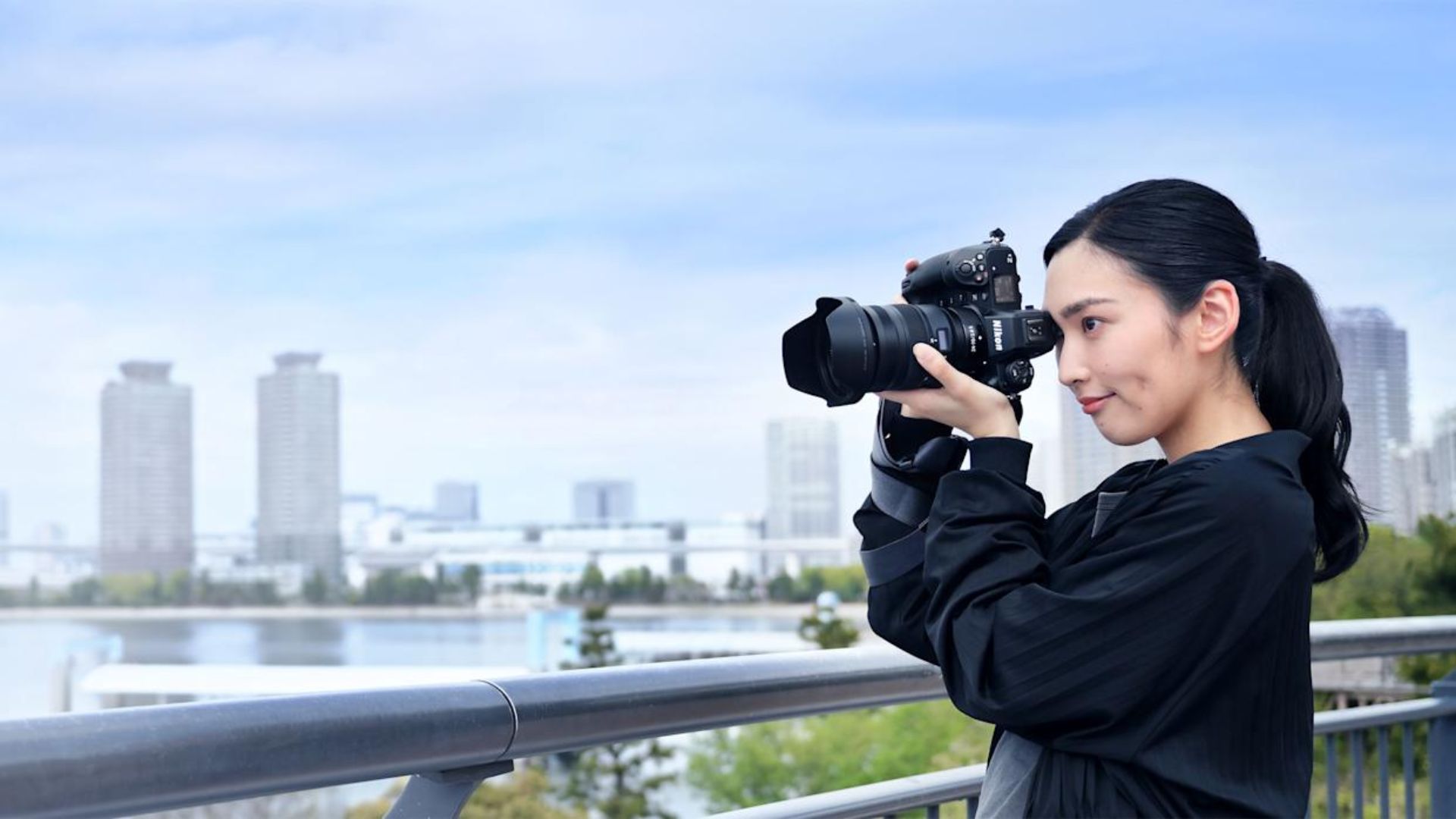 Asian woman in black outfit with ponytail holding DSLR camera photographing urban skyline with tall buildings across waterfront from elevated viewpoint with metal railing.