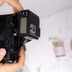 Person holds black DSLR camera showing LCD screen and controls while shooting product photos with blurred props in bright background.