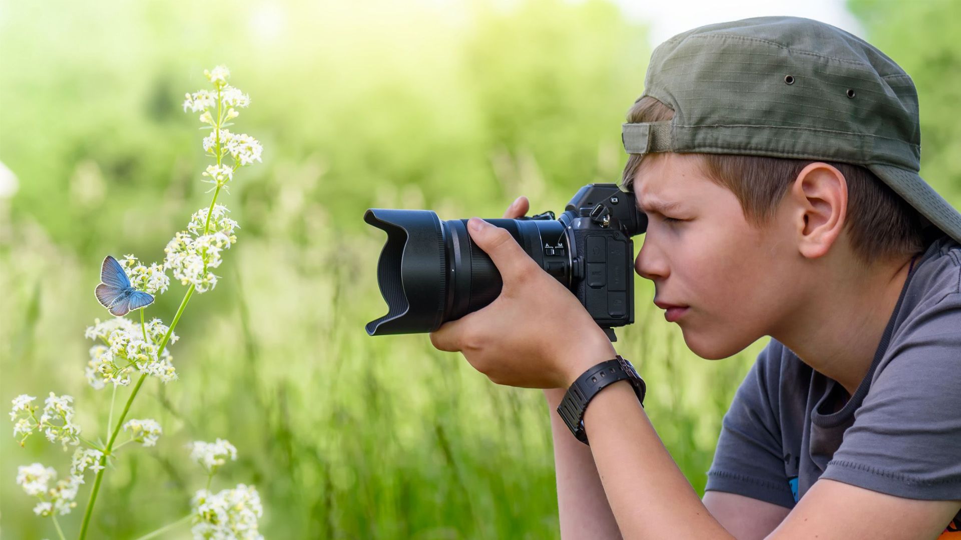 Child wearing olive green cap uses mirrorless camera to capture blue butterfly resting on white wildflowers in sunny meadow setting.