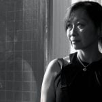 Black and white portrait of mature Asian woman wearing sleeveless black top and necklace standing thoughtfully by window with urban view.