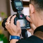 Wedding photographer in dark formal attire holding DSLR camera with external flash unit mounted on top reviewing images on LCD screen indoors.