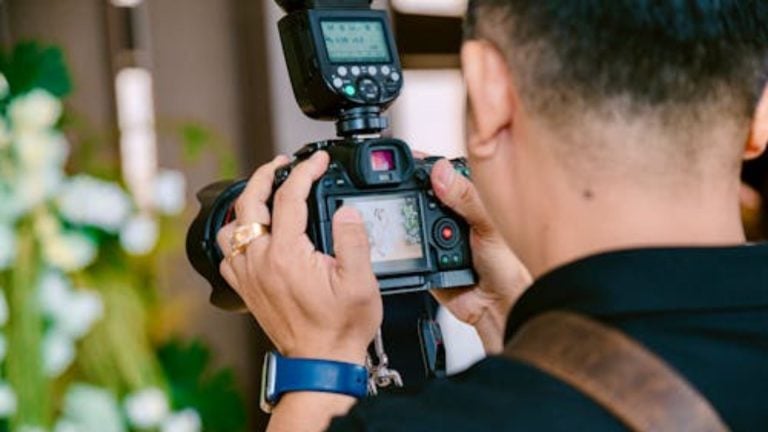 Wedding photographer in dark formal attire holding DSLR camera with external flash unit mounted on top reviewing images on LCD screen indoors.