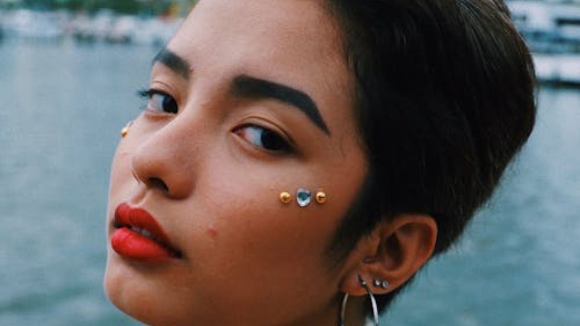 Close-up of woman with short dark hair, bold red lipstick, multiple facial piercings including medusa and nostril studs, ear piercings against blurred background.