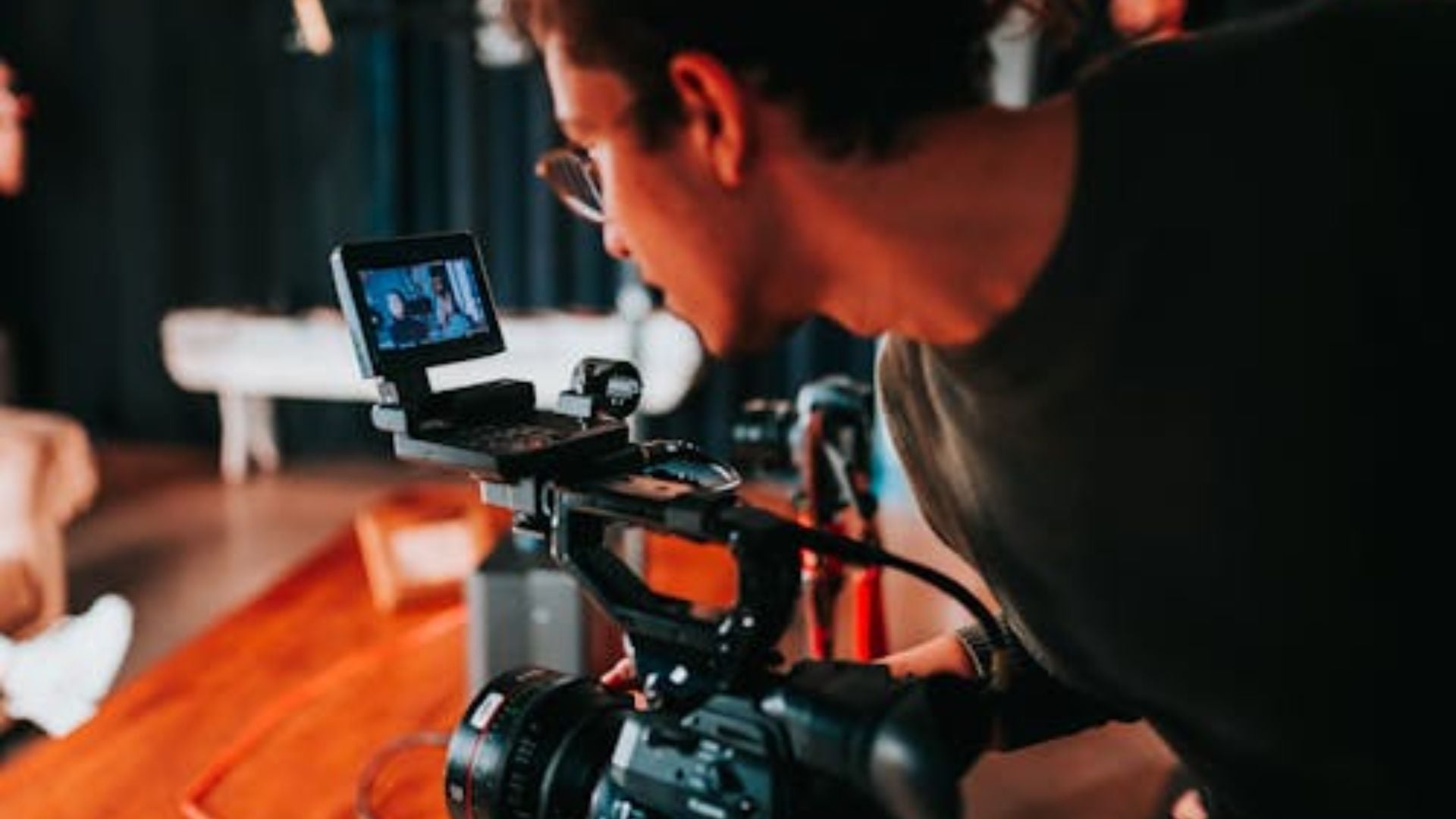 Person operating professional video camera with mounted monitor screen during indoor production shoot, focused on capturing content with stabilized rig and orange lighting.
