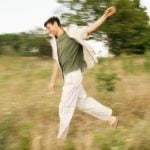 Man in green shirt and white pants captured mid-stride running through grassy field with motion blur effect and trees in background.