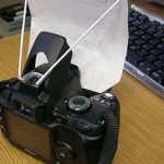 Black digital camera on bamboo mat features white pop-up flash diffuser beside keyboard showing DIY photography lighting modification for better photos.