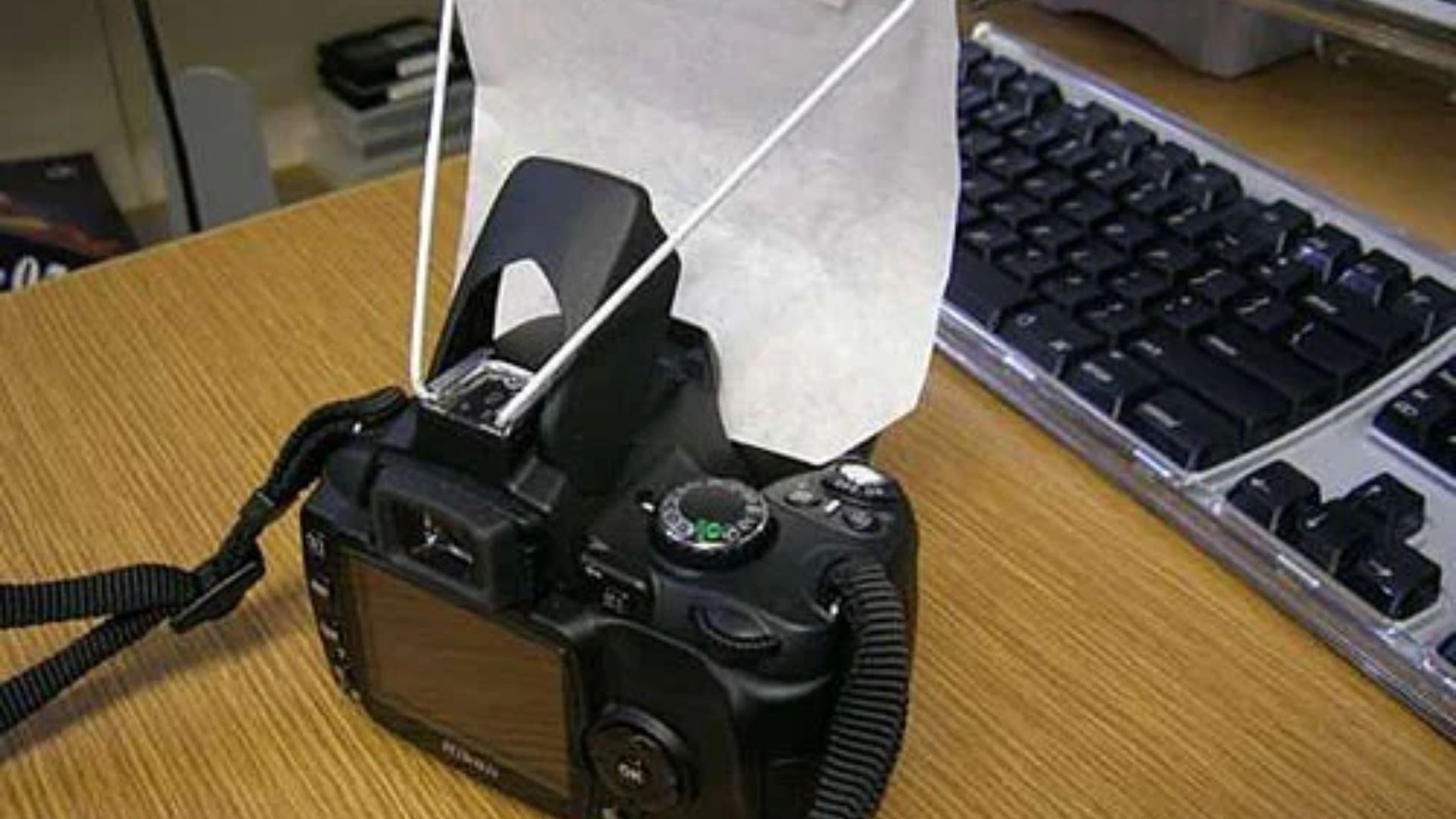 Black digital camera on bamboo mat features white pop-up flash diffuser beside keyboard showing DIY photography lighting modification for better photos.