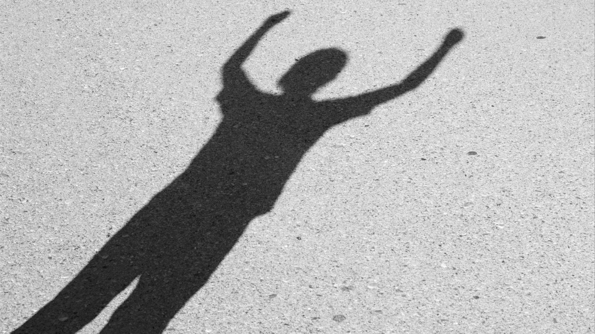 Dark shadow cast on light concrete ground showing figure with both arms raised up in celebratory or joyful gesture overhead.