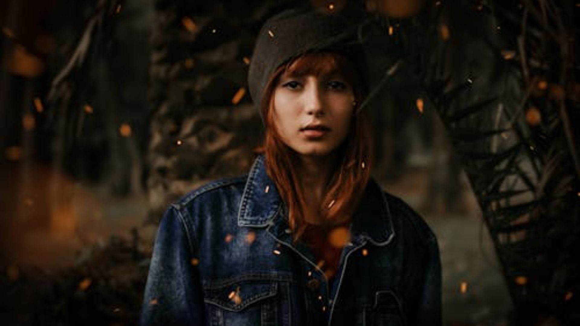Woman with long auburn hair wearing gray cap and blue denim jacket outdoors with floating golden particles, bare branches visible in moody atmospheric background.