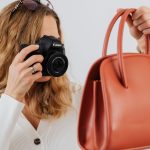 Blonde woman with sunglasses on head holding Canon DSLR camera photographing coral orange structured handbag against white wall wearing white ribbed cardigan.