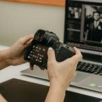 Hands holding digital camera showing LCD screen with camera settings while reviewing photos at white desk with laptop displaying wedding photos in background workspace.